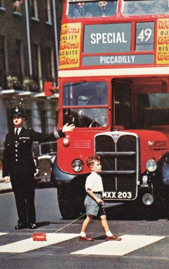 პატარა ბიჭი კვეთს ქუჩას, ხელში თოკზე მობმული სათამაშო ავტობუსით. ლონდონი (1960წ)